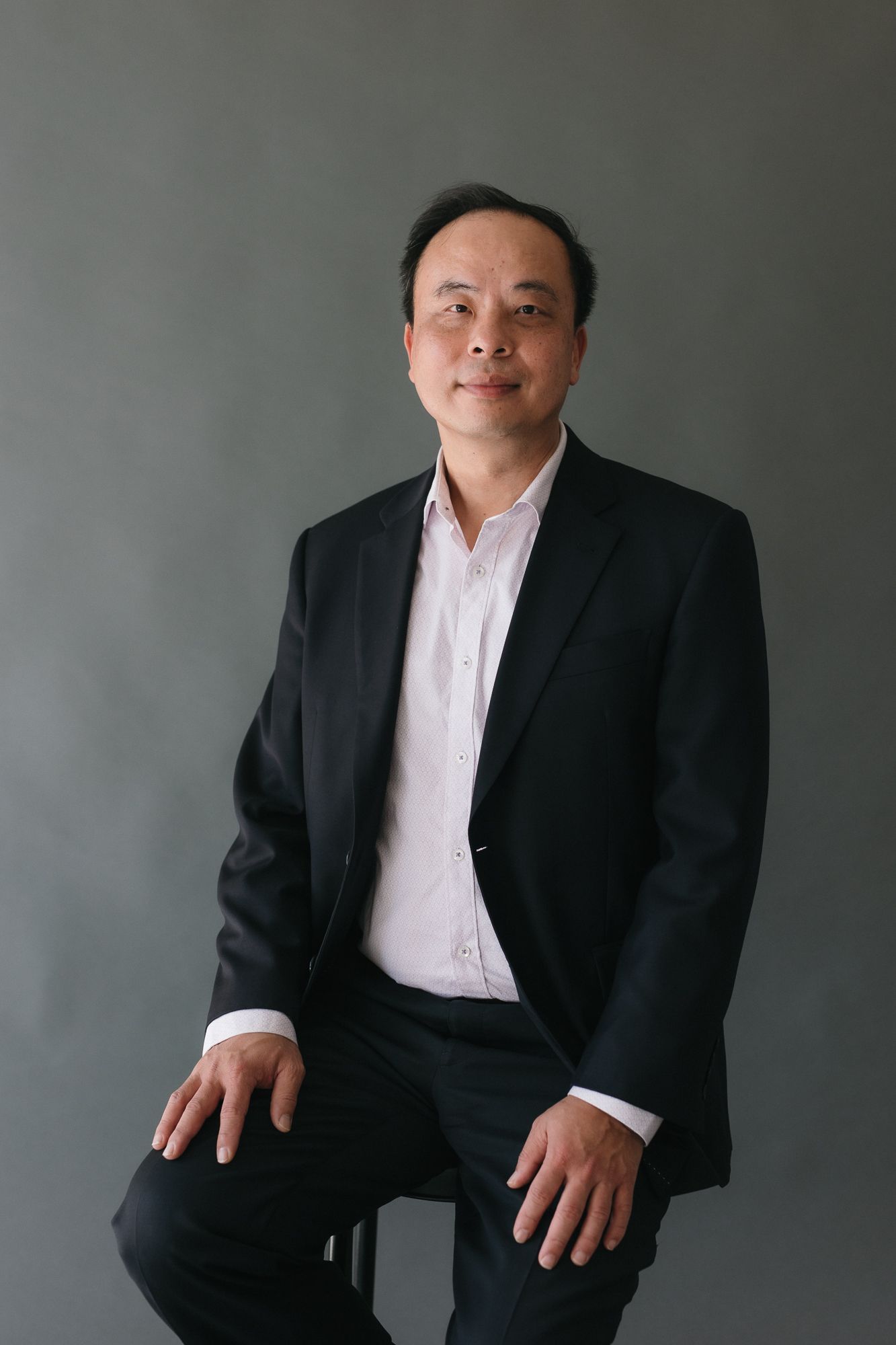 Middle-aged Asian man in black blazer and white shirt seated confidently against a gray studio background.
