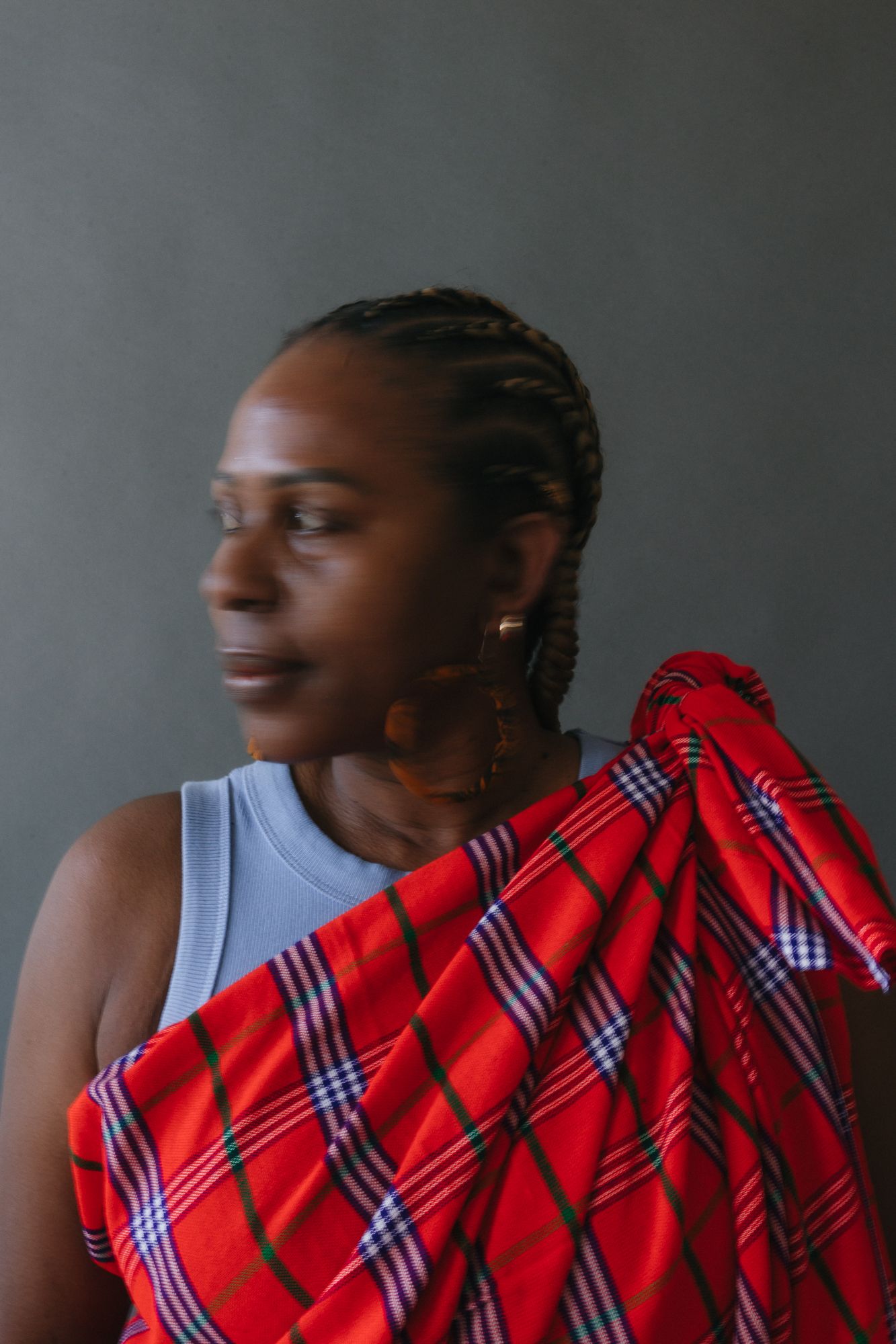A Black woman with braids wears a gray tank top and a red plaid shuka draped over her shoulder, looking sideways.