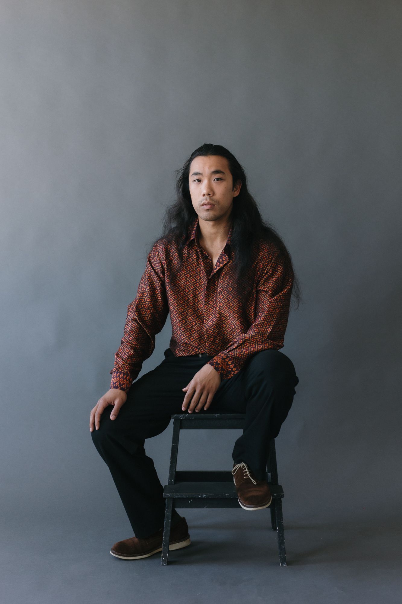 Young Asian man with long hair sits on a stool wearing a red patterned shirt and black pants against gray backdrop.