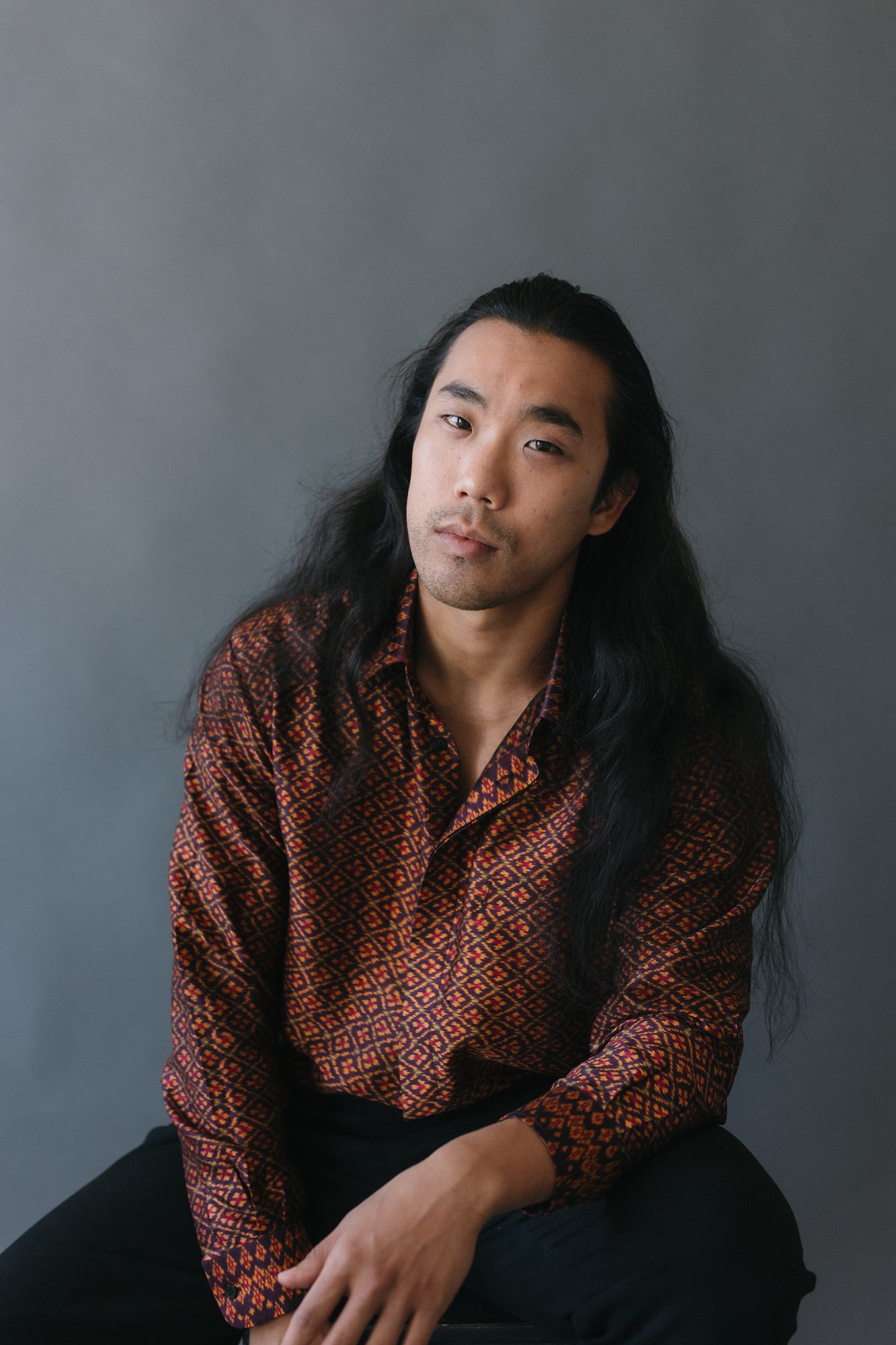 Young Asian man with long black hair wearing a patterned red shirt, seated against a gray background.