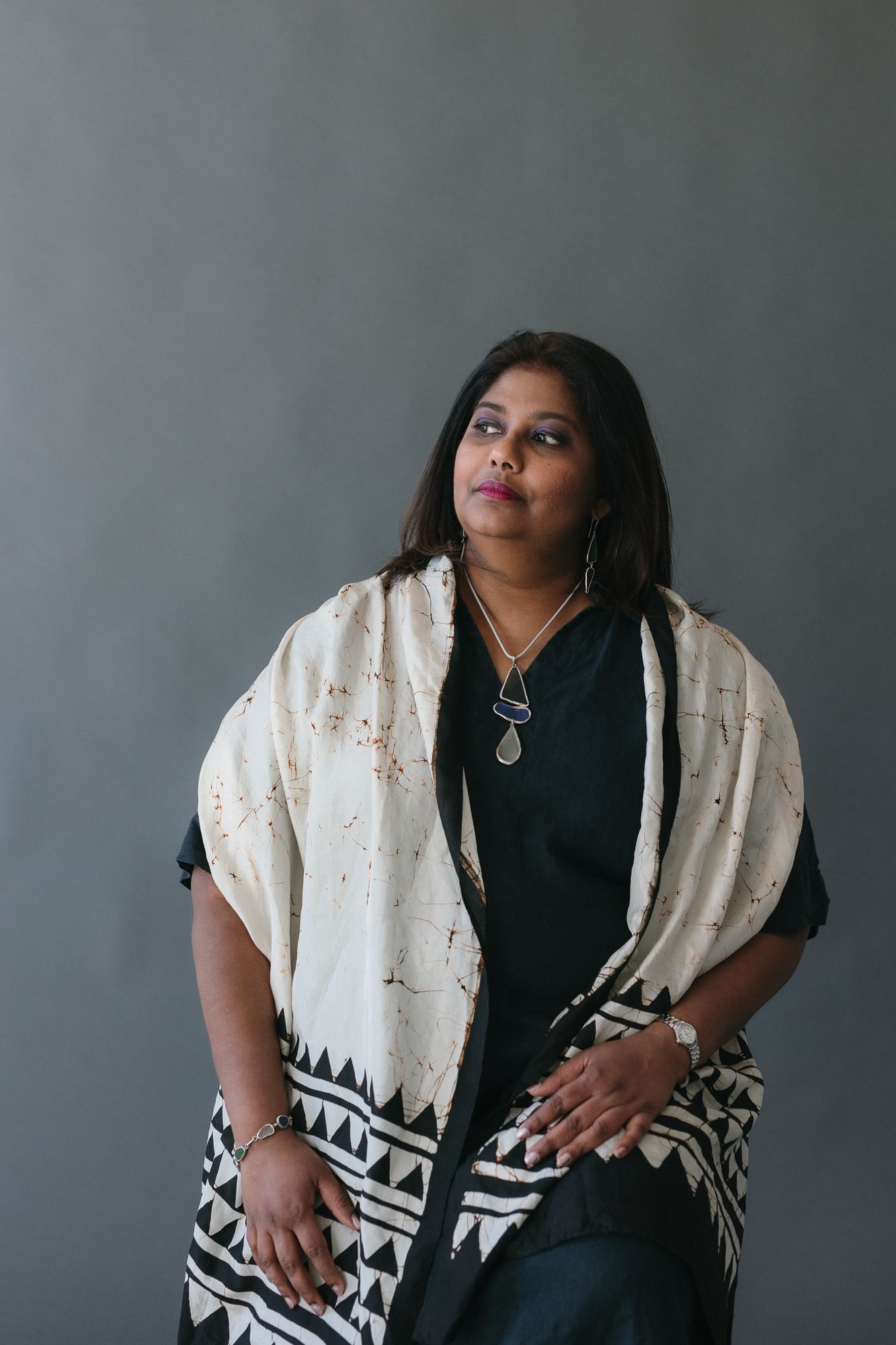 A woman in black outfit with white geometric patterned shawl and silver jewelry poses against gray background.