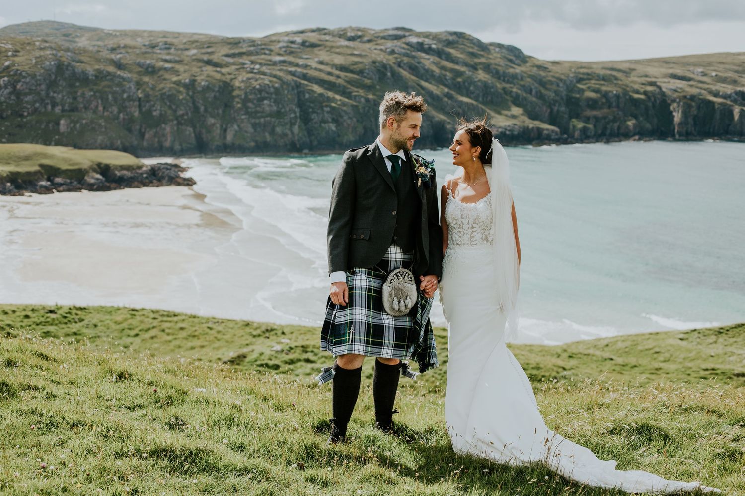 Couple in Scottish tartan kilt and white wedding gown hold hands overlooking a scenic coastal beach landscape.