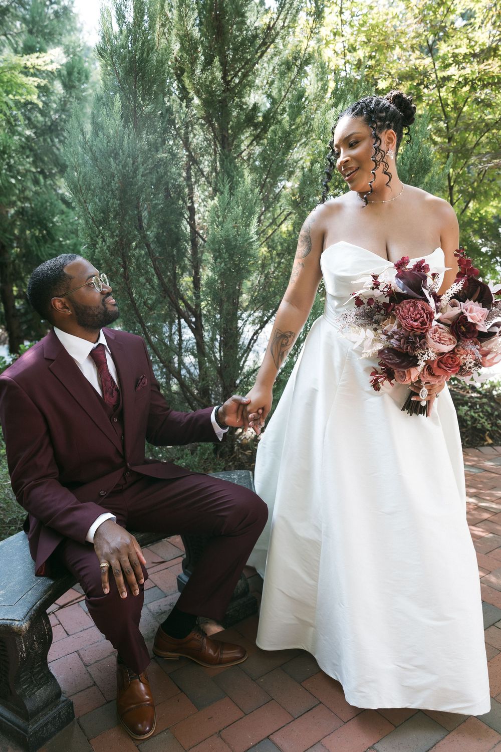 A groom in a burgundy suit holds hands with his bride in a white strapless gown holding a mauve bouquet outdoors.