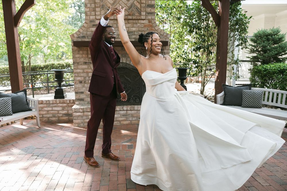 Joyful newlyweds celebrate outdoors, bride in white gown twirling as groom in burgundy suit raises arms in triumph.