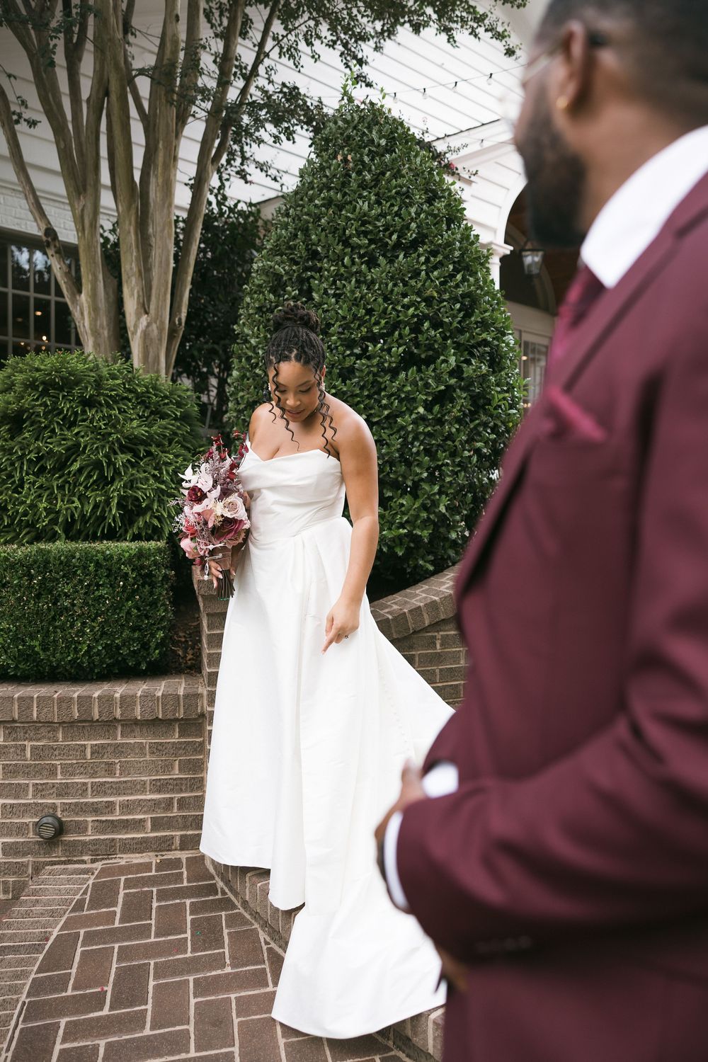 Bride in white strapless gown holds floral bouquet on brick steps while groom in burgundy suit stands in foreground.