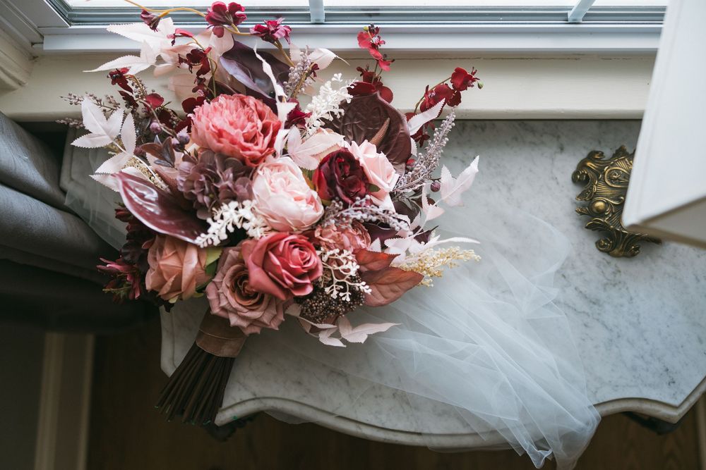 Romantic bridal bouquet with dusty pink roses, burgundy foliage, and white blooms resting on a marble surface.