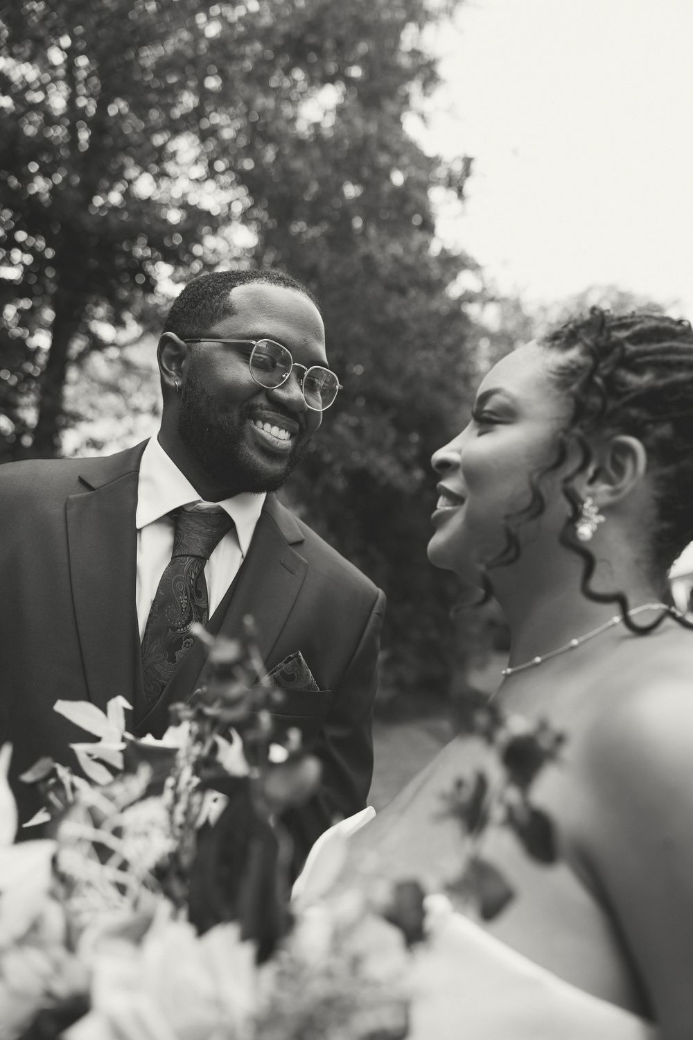 Black and white photo of a smiling groom in suit and glasses gazing at his bride holding a bouquet outdoors.