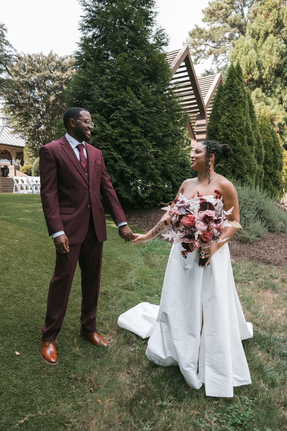 A groom in a burgundy suit holds hands with his bride in a white gown holding a floral bouquet in a lush garden.