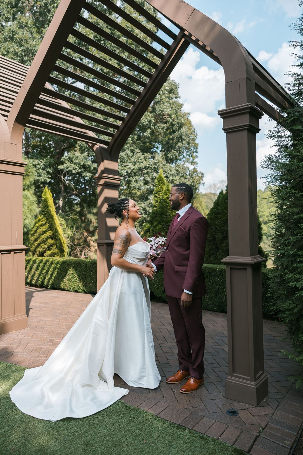 A bride and groom share a romantic moment under a wooden pergola arch surrounded by lush greenery on their wedding day.