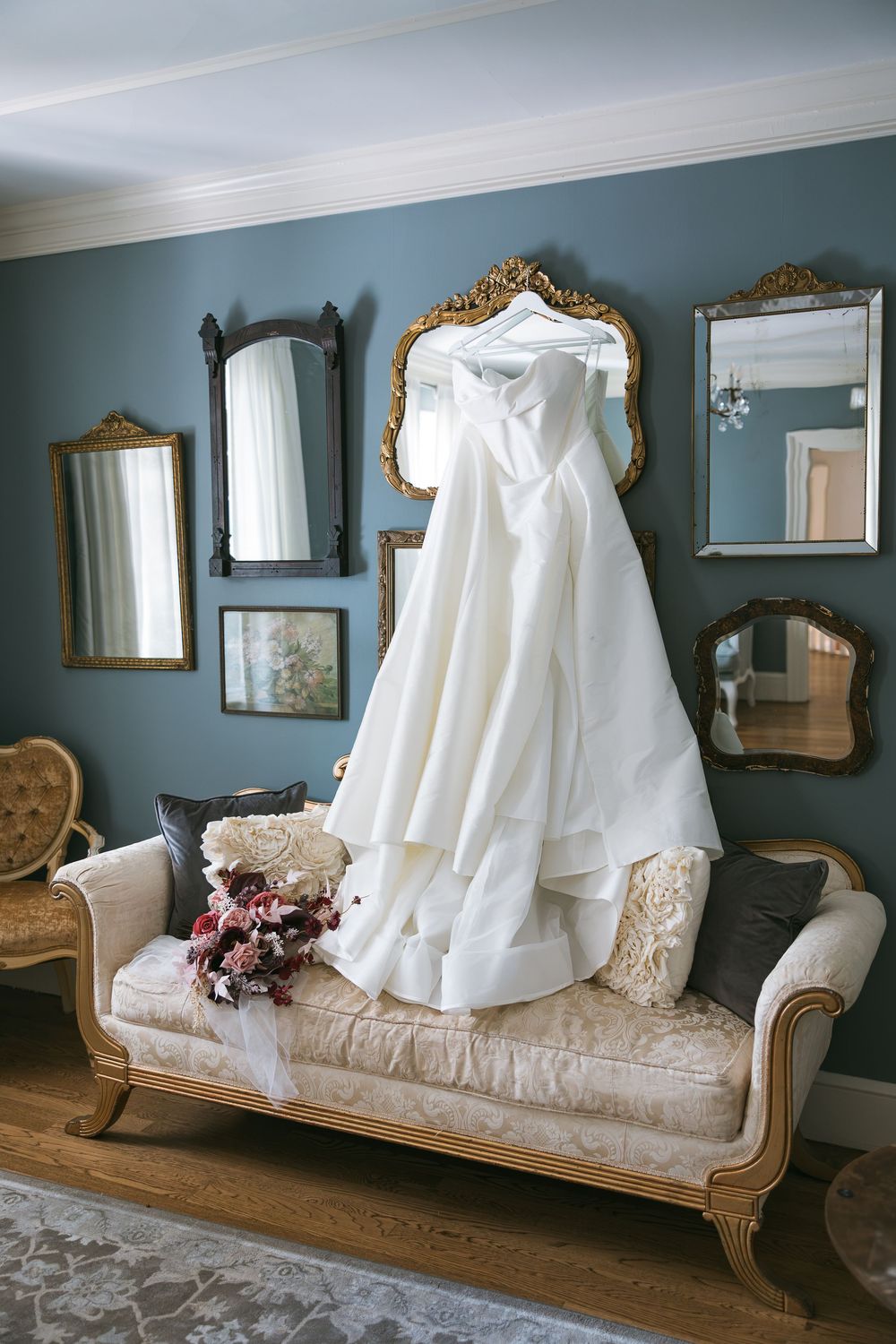 Elegant white wedding dress hanging on vintage chaise lounge surrounded by ornate gold mirrors on blue-gray wall.