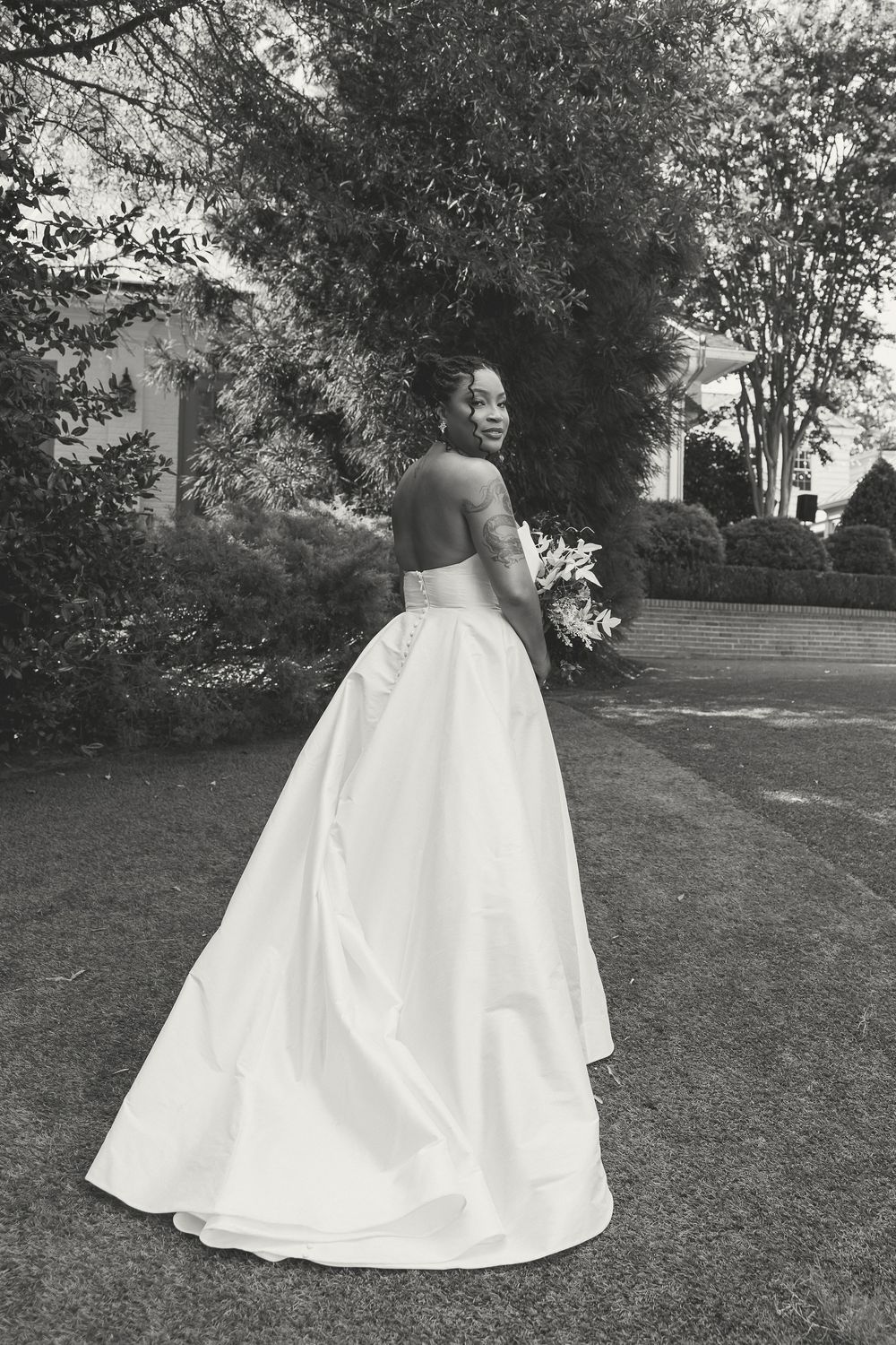 Black and white photo of a bride in a strapless ballgown holding a bouquet, posing in a lush garden setting.