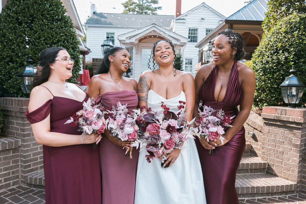 Bride in white gown laughs with three bridesmaids in mauve dresses holding floral bouquets outside brick venue.