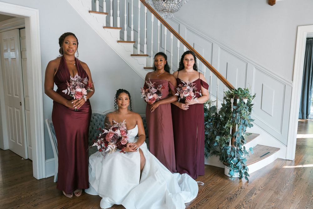 Bride in white gown seated with three bridesmaids in burgundy dresses holding bouquets near elegant staircase.