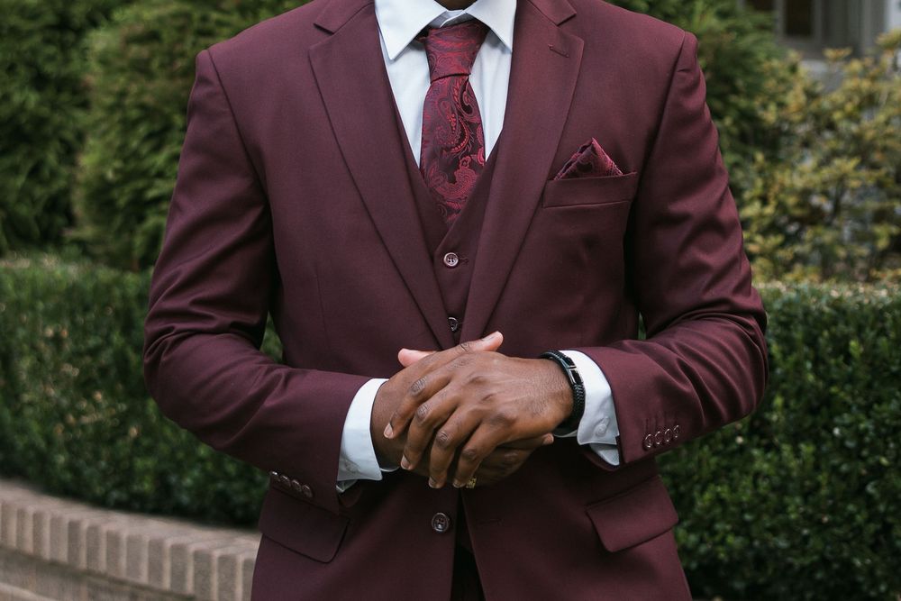 Man in burgundy three-piece suit with matching tie and pocket square, hands clasped, standing outdoors.