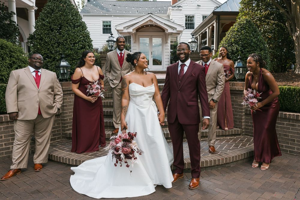 Wedding party poses outside a brick venue, bride in white gown, groom in burgundy suit, bridesmaids in wine dresses.