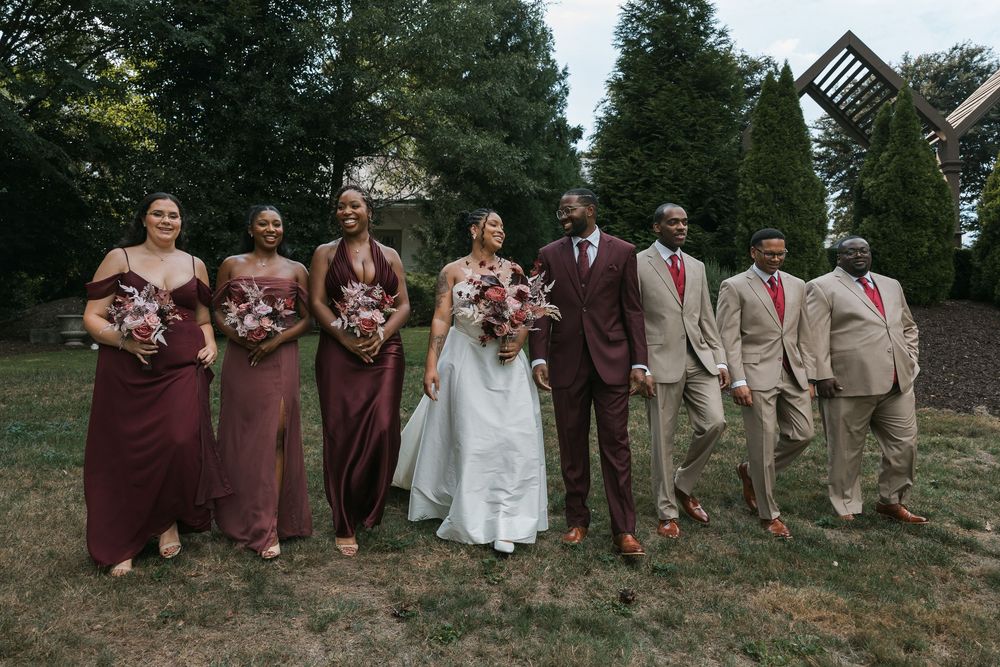 Wedding party poses outdoors, bride in white gown with bridesmaids in burgundy dresses and groomsmen in tan suits.