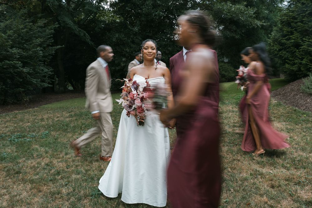 Bride in white gown holding pink bouquet stands with wedding party in burgundy dresses on green lawn.