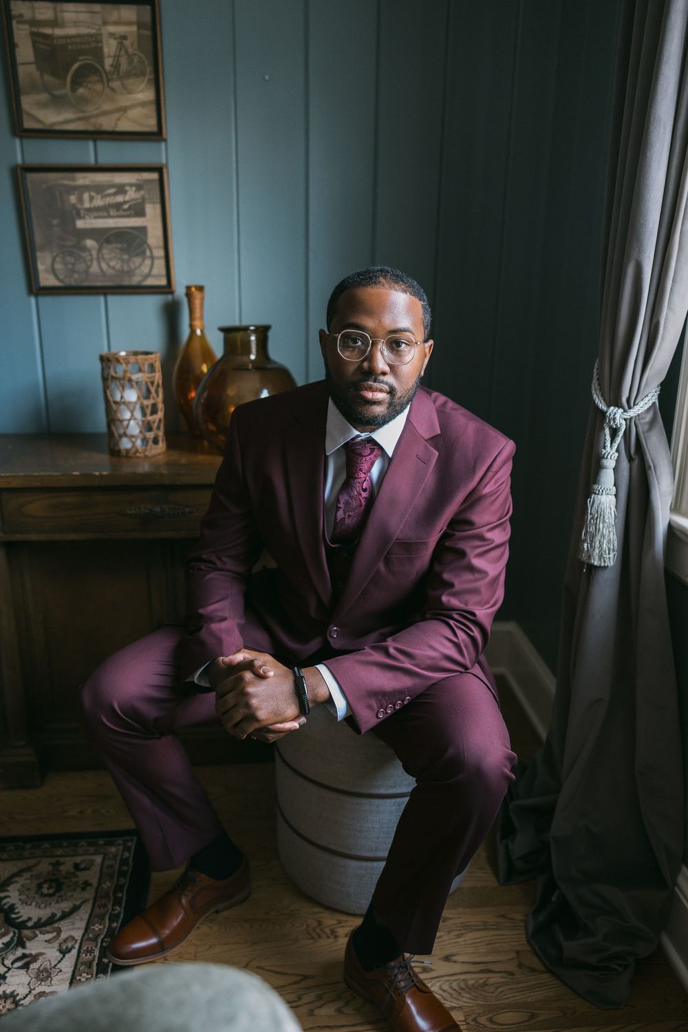 A well-dressed Black man in a burgundy suit and glasses sits confidently on a stool in a stylish vintage-inspired room.