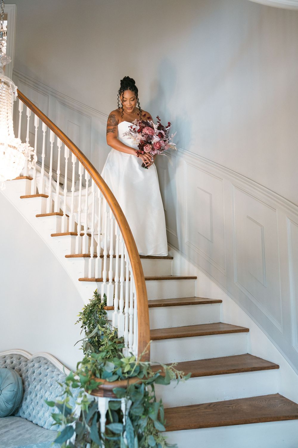 Bride in white gown descends elegant curved staircase holding pink floral bouquet in bright white interior.