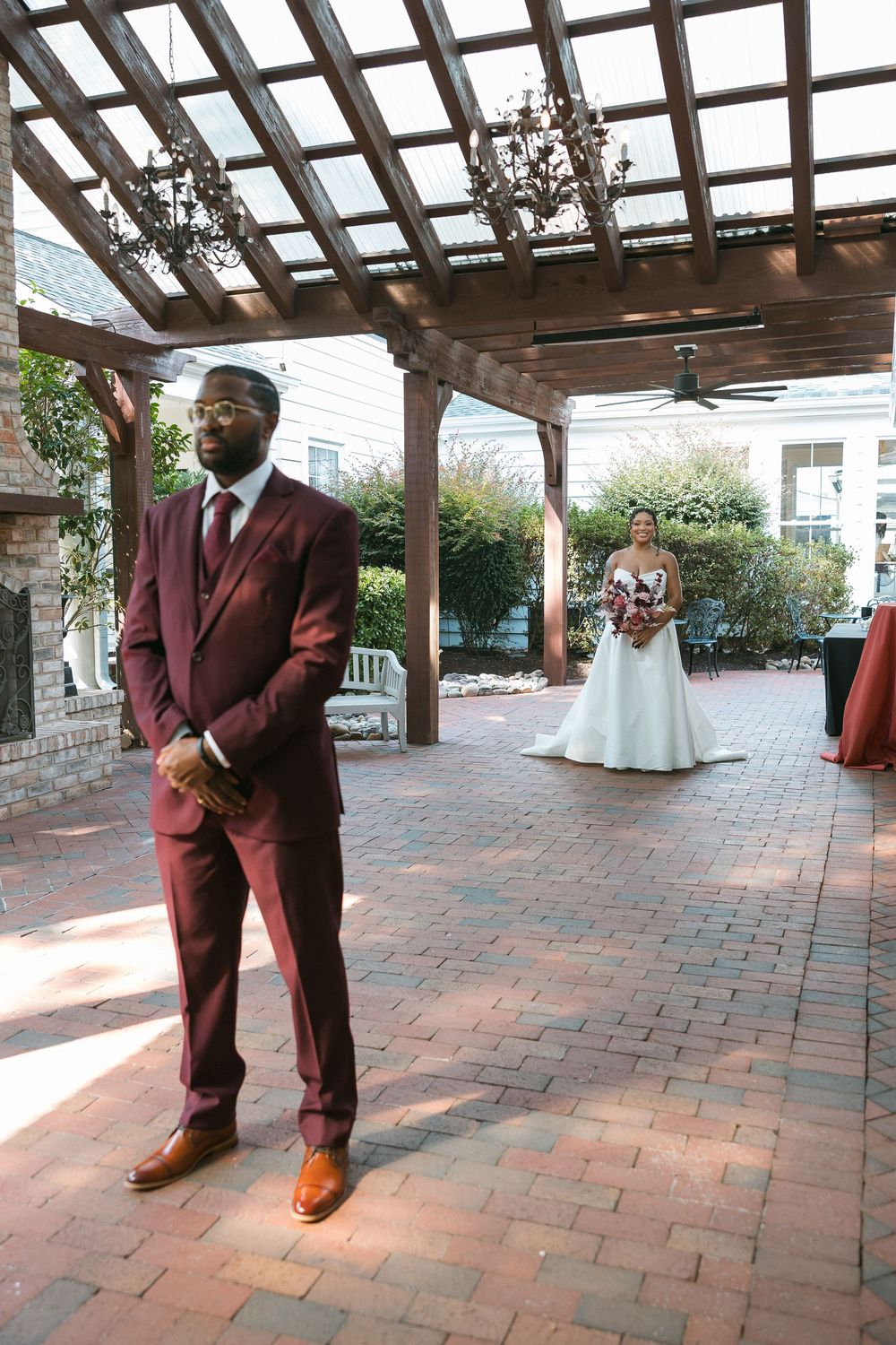Groom in burgundy suit awaits bride in white gown during first look moment on brick patio under wooden pergola.