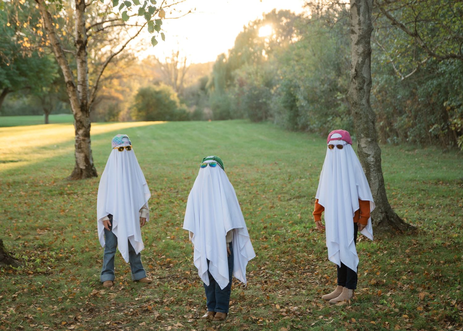 Three children in white sheet ghost costumes with sunglasses and hats stand on a grassy lawn among trees at golden sunset.