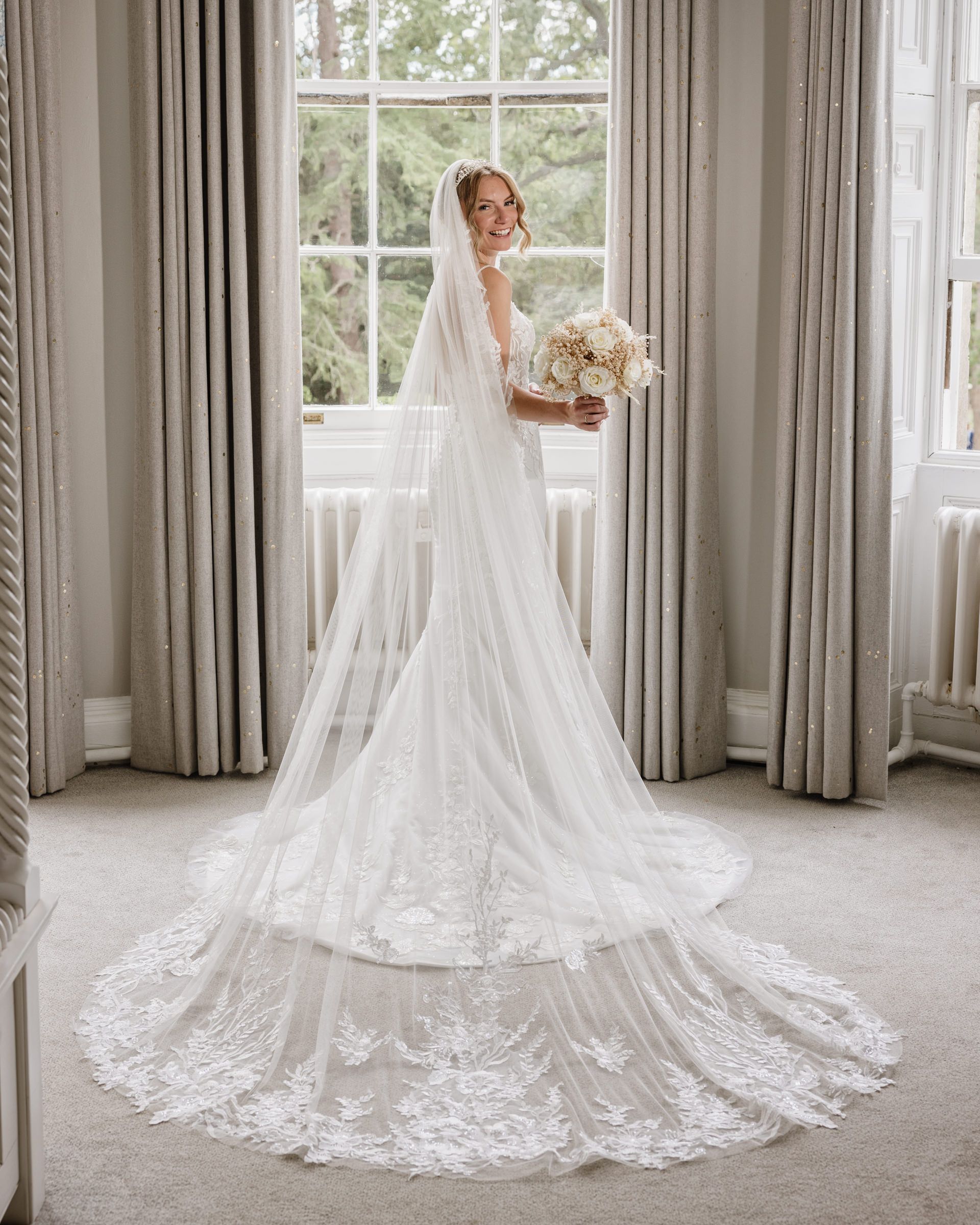 Smiling bride in a lace wedding gown with a long cathedral veil holding a white bouquet beside a tall window in an elegant room.