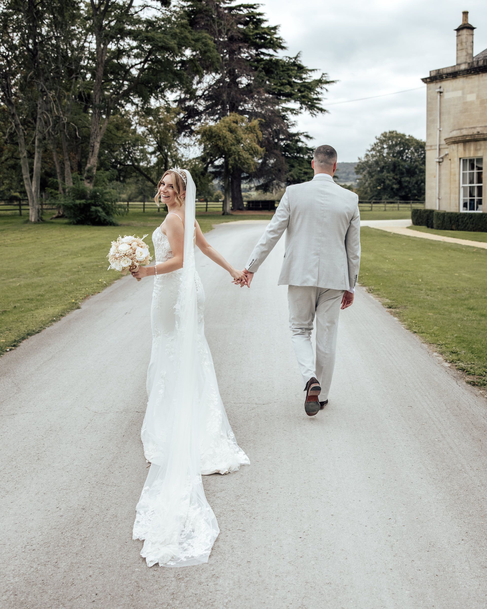 Bride in a lace wedding gown with a long veil holds a bouquet and walks hand in hand with her groom down a driveway toward a country manor.