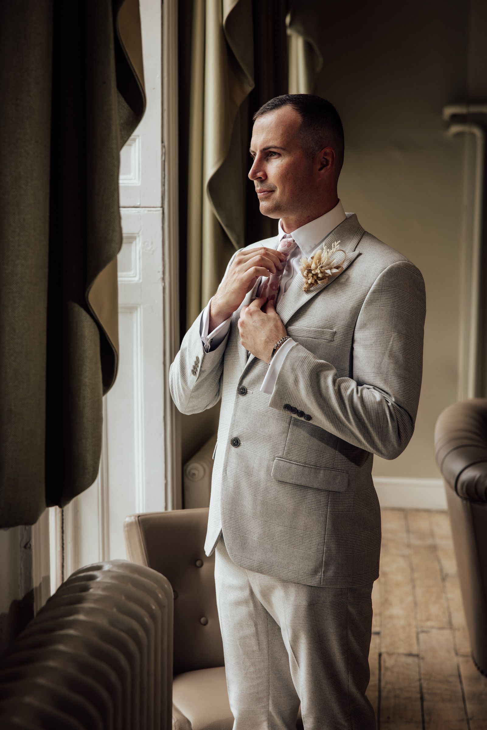 Groom in a light gray suit with a floral boutonniere adjusts his tie while gazing out a window in an elegant room with wood floors.