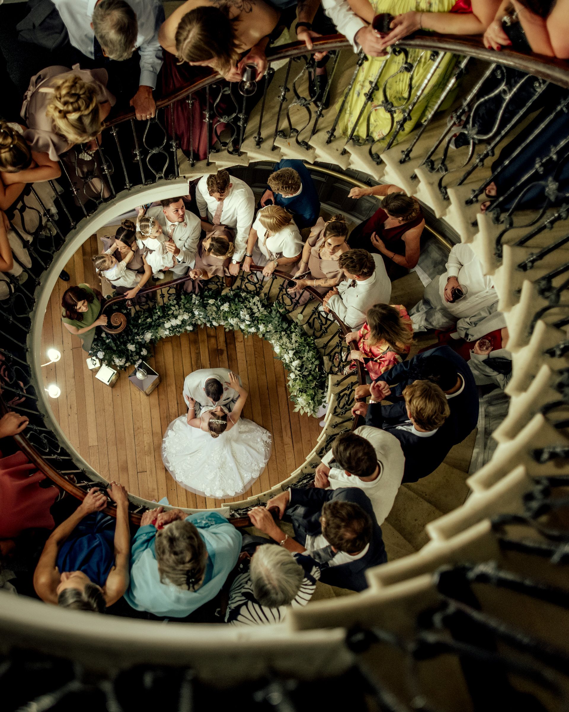 Overhead view of a bride and groom dancing on a wooden floor as wedding guests watch from a curved staircase.