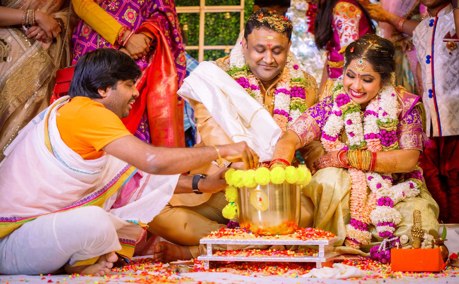 Bride and groom in floral garlands and traditional attire perform a wedding ritual with a priest and brass pot during a Hindu ceremony.