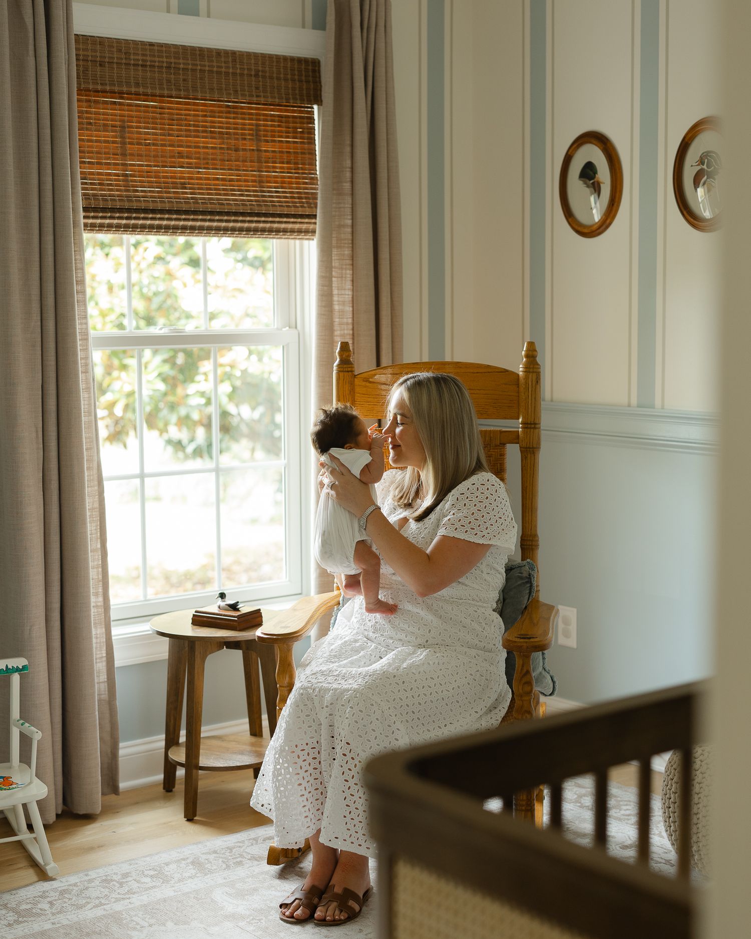 A mother in a white eyelet dress holds her newborn baby while seated in a wooden rocking chair beside a sunlit window in a cozy nursery.