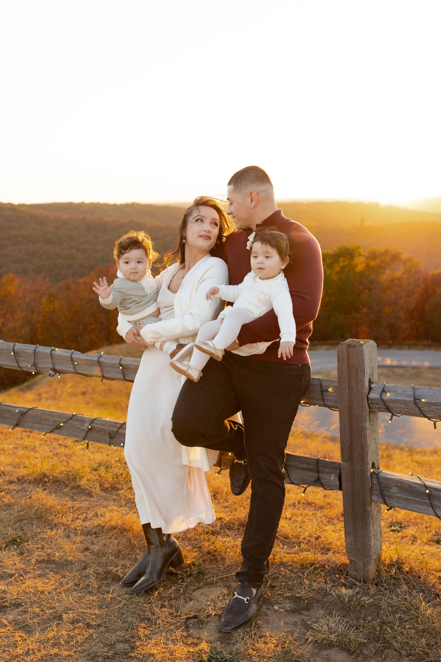 Family of four posing under vibrant red autumn tree, parents with two young children on a sunny fall day.