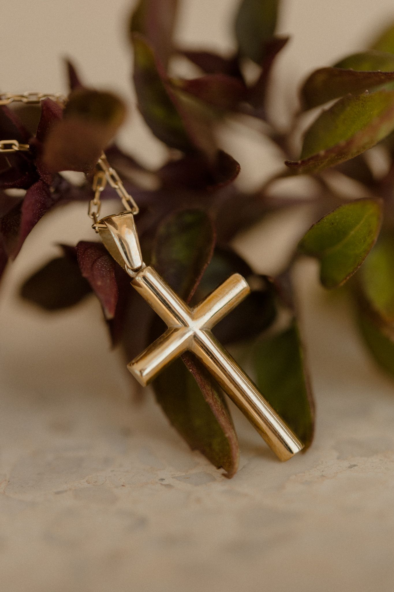 Gold cross pendant necklace leaning against dark green and burgundy leaves on a marble surface.