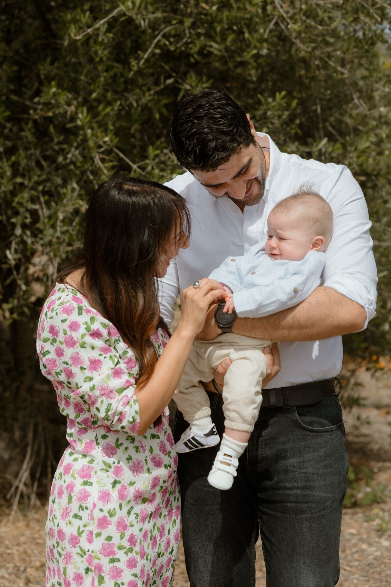 A young family outdoors, with a man in a white shirt holding a baby while a woman in a floral dress tenderly interacts with the child.