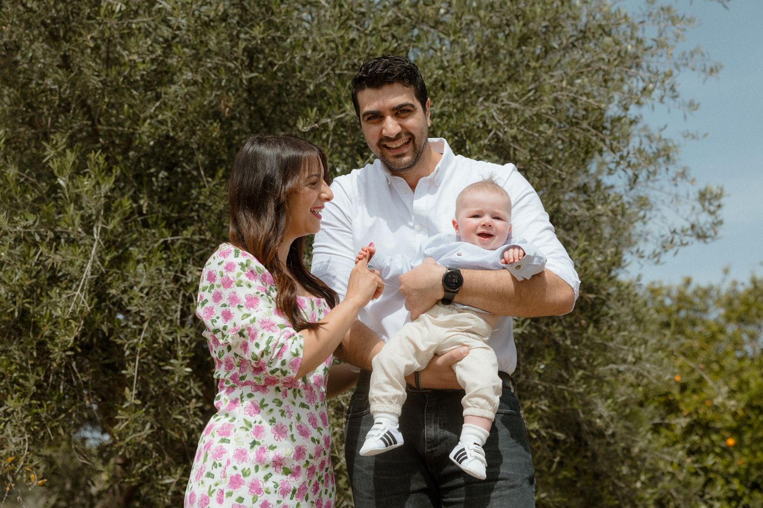 Happy young couple smiling and holding their baby outdoors among green trees on a sunny day.