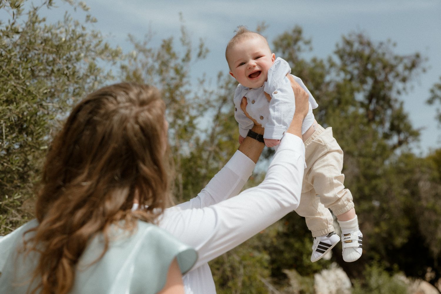 A joyful mother lifts her laughing baby into the air outdoors, surrounded by green trees on a bright day.