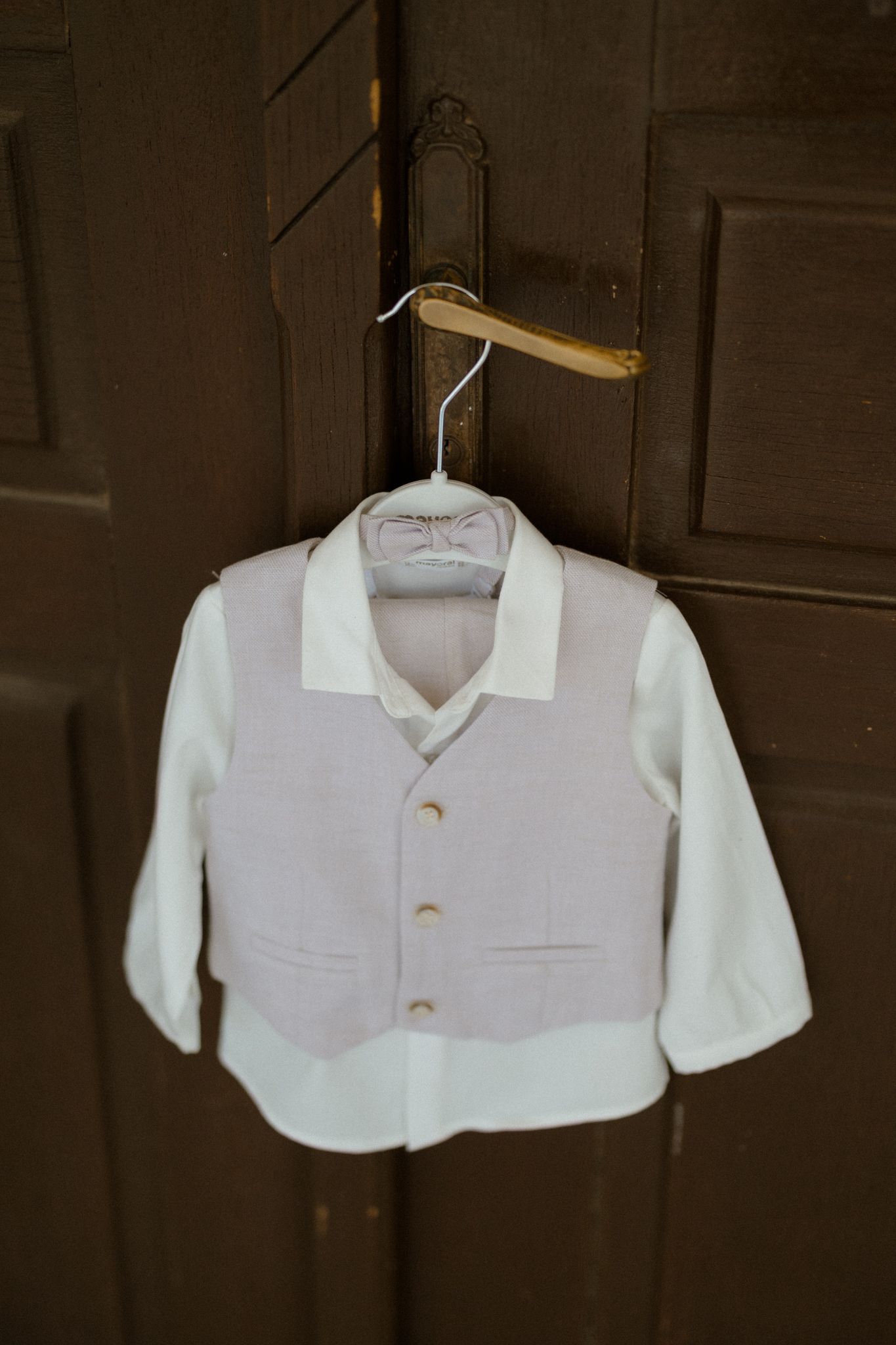 Small boys white dress shirt and gray vest outfit on a wooden hanger hung on a dark wood door.