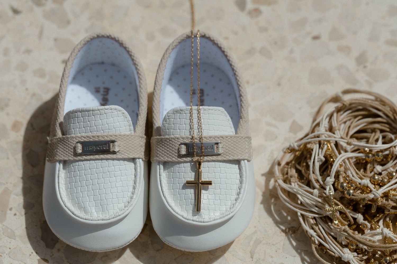 White baby loafer shoes with a gold cross charm and tan strap, placed on a light stone surface.