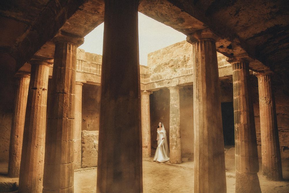 Bride in white gown standing among ancient stone columns in historic ruins.