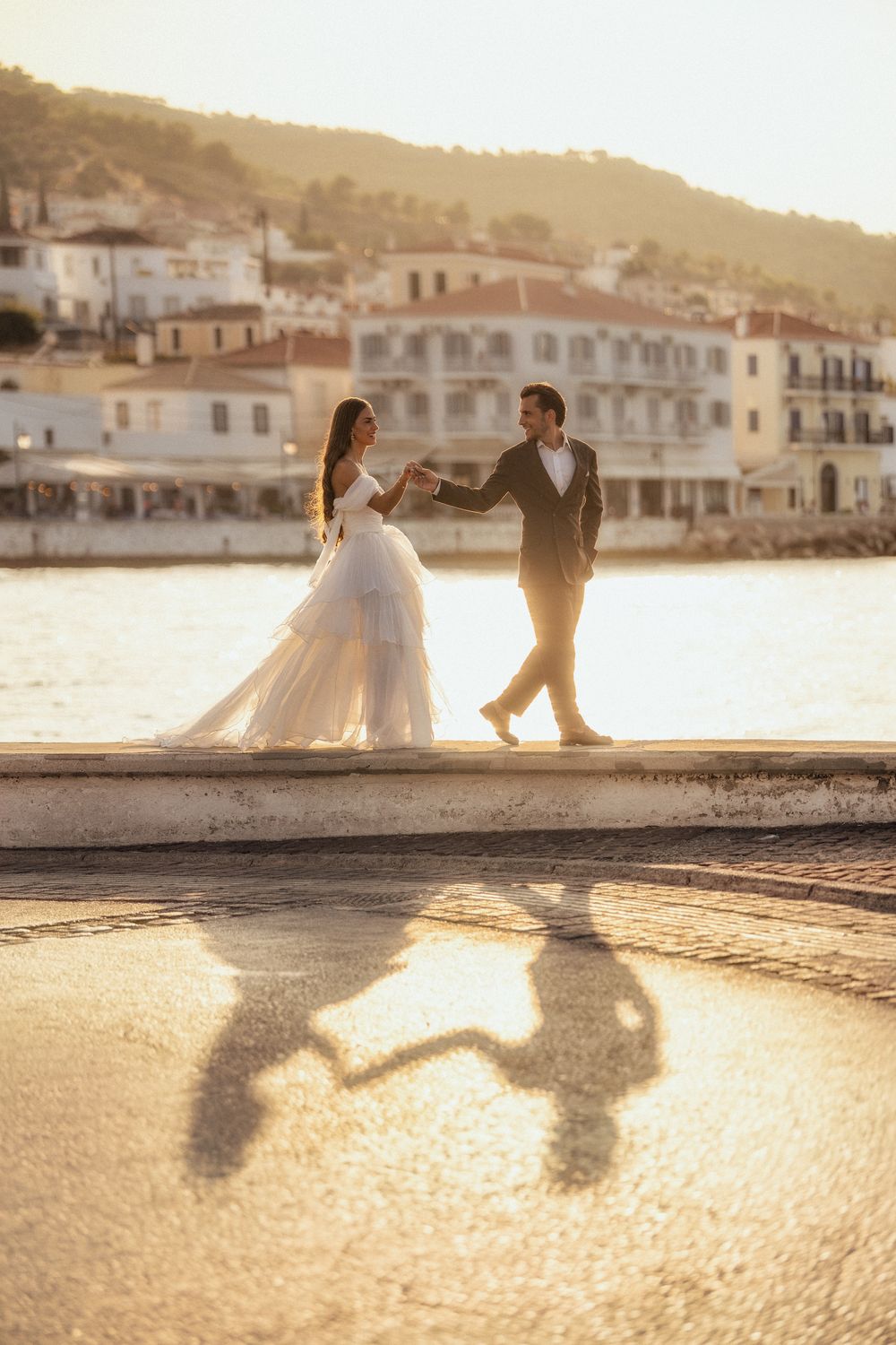 Bride and groom dancing on a waterfront promenade at golden hour with Mediterranean village backdrop.