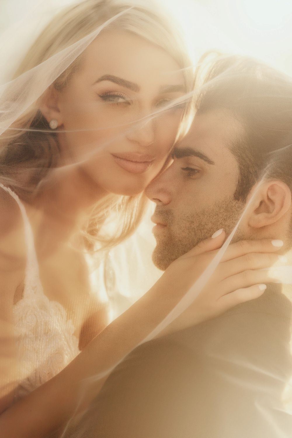 Romantic couple almost kissing under a bridal veil in soft golden light, bride in lace touching groom's face.