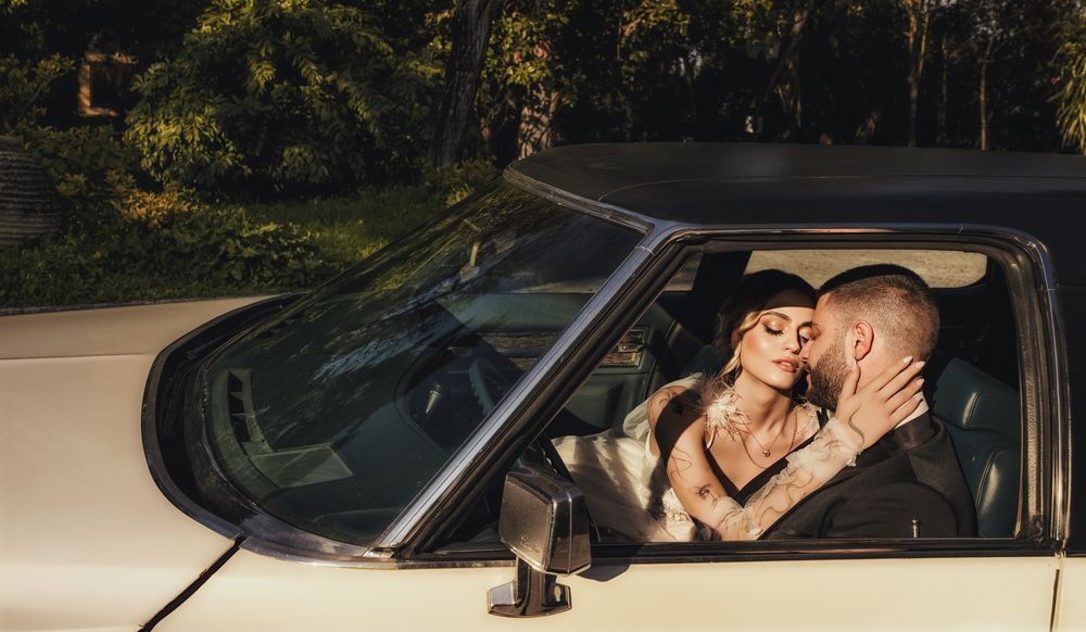 A tattooed couple shares a romantic kiss inside a classic cream convertible car at night.