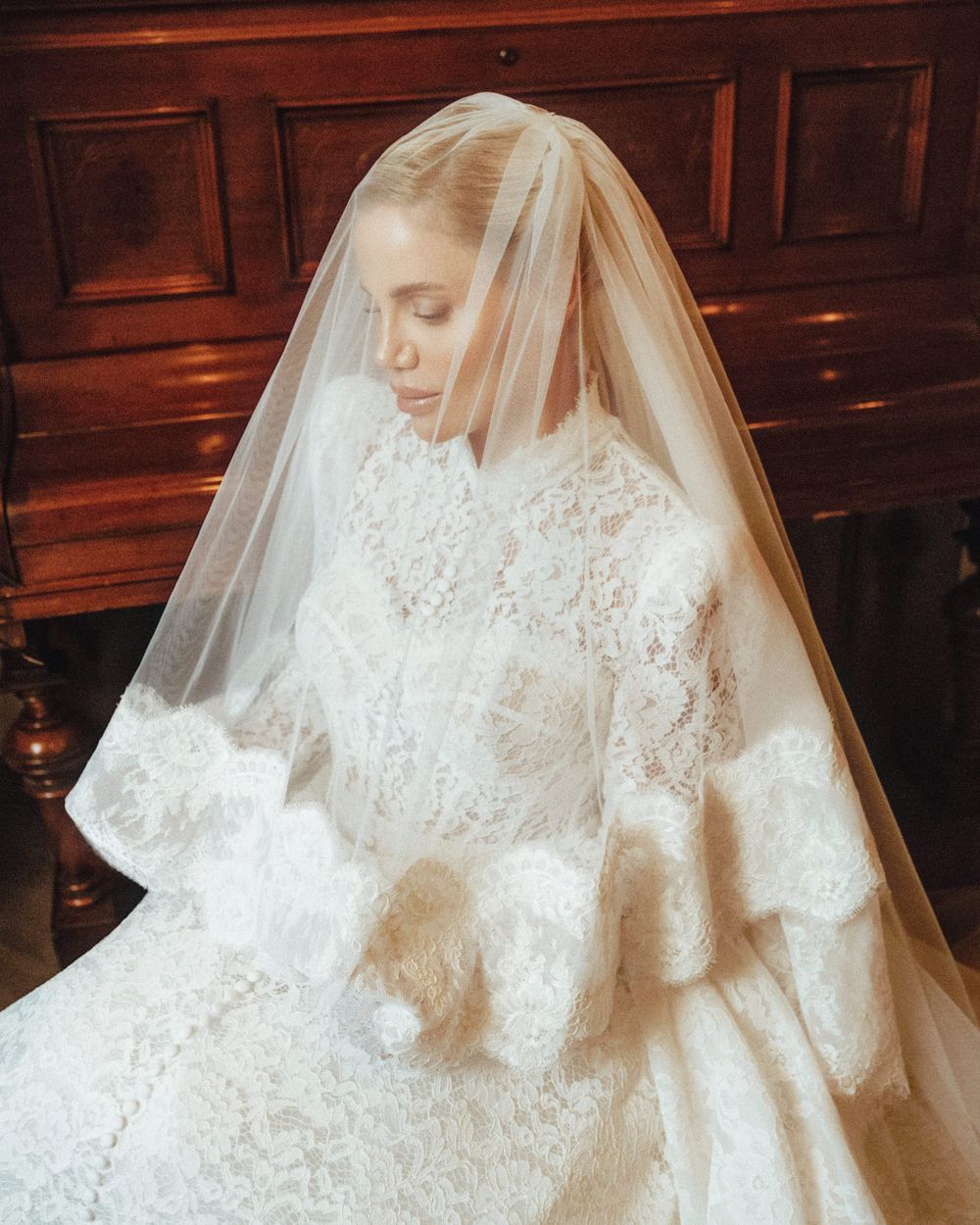 Bride in elegant long-sleeve lace wedding gown with flowing veil, posing gracefully in a warmly lit wooden interior.