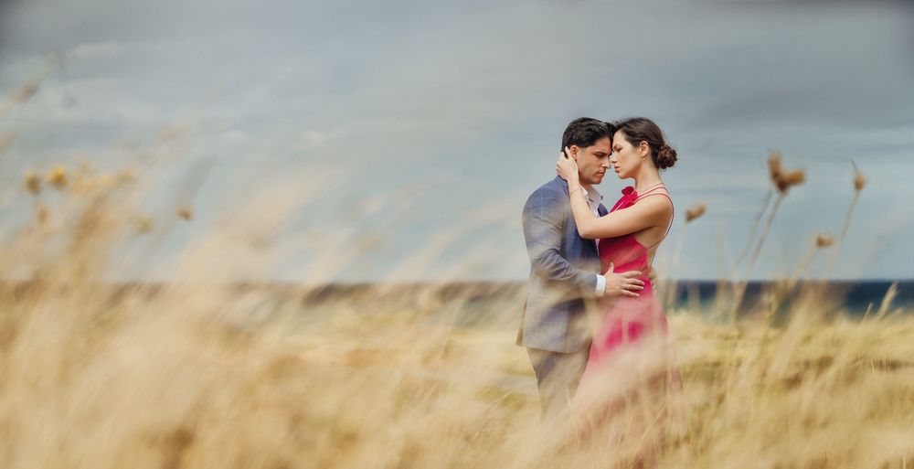 Romantic couple embracing in golden wheat field with dramatic cloudy sky backdrop at seaside location.