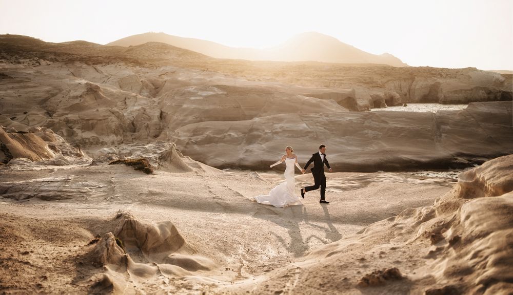 Couple in wedding attire running through golden desert landscape at sunset.
