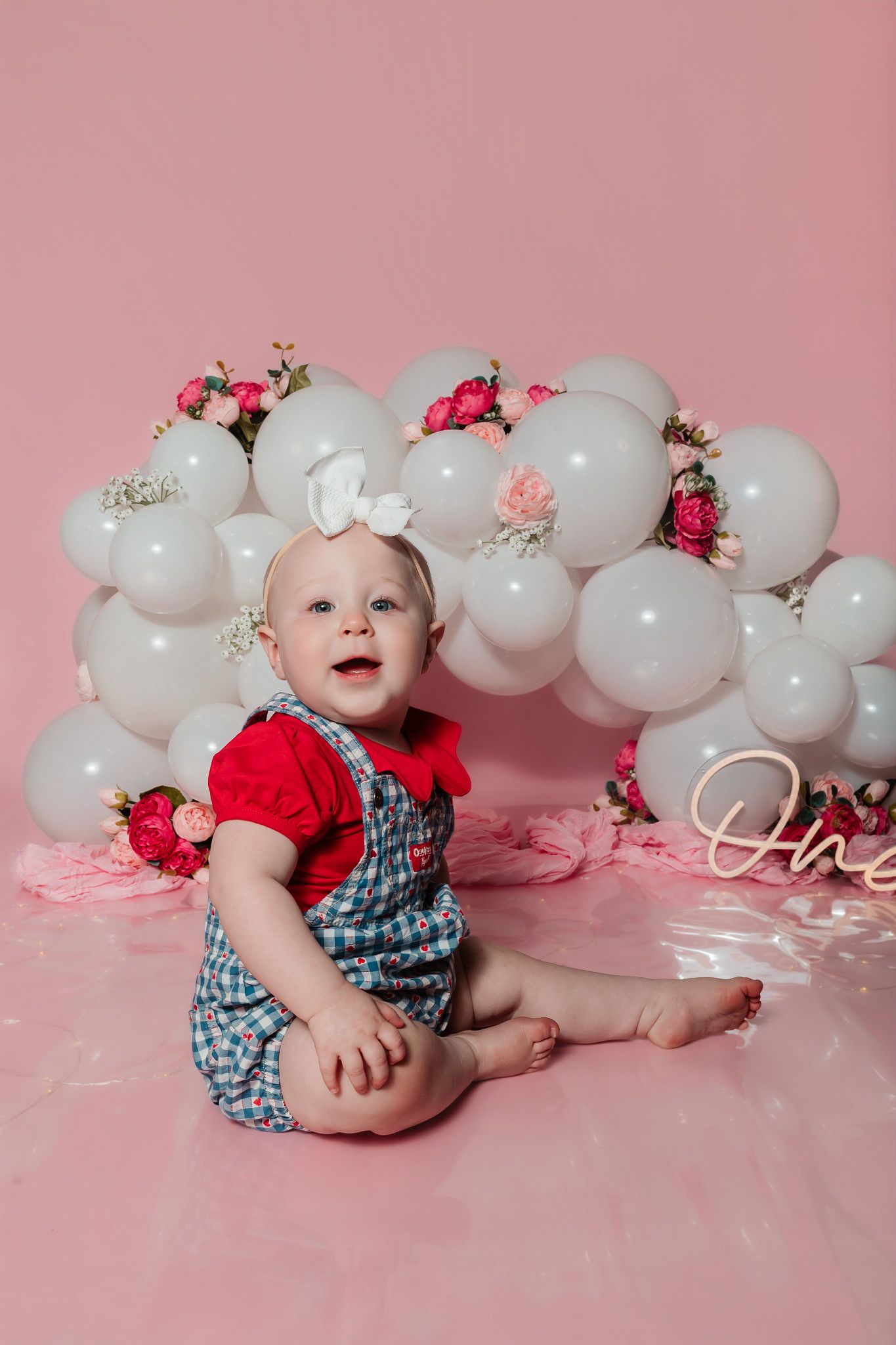 Baby girl in red top and overalls sits before white balloon arch with flowers for first birthday cake smash photo.