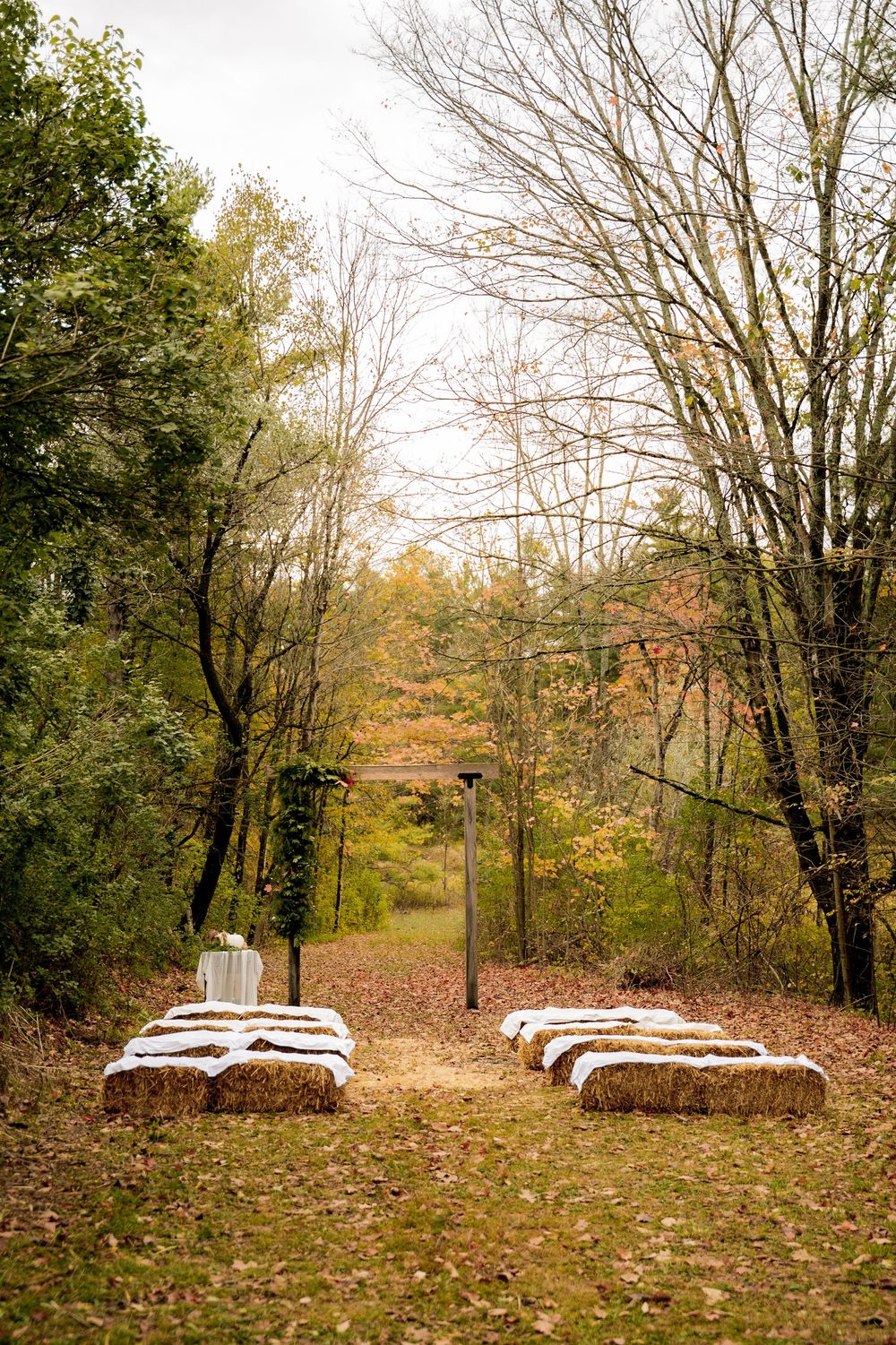 Rustic outdoor wedding ceremony setup with hay bale seating along a forest aisle in autumn.