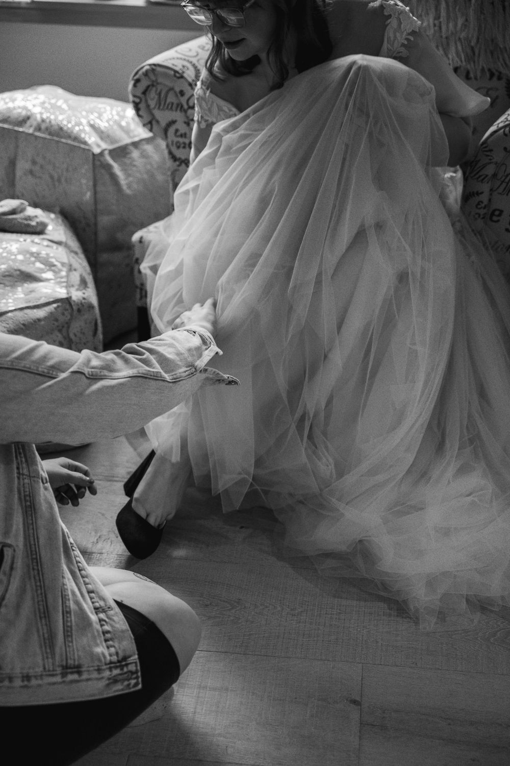 Black and white photo of bride in tulle gown getting shoes put on by bridesmaid before wedding ceremony.