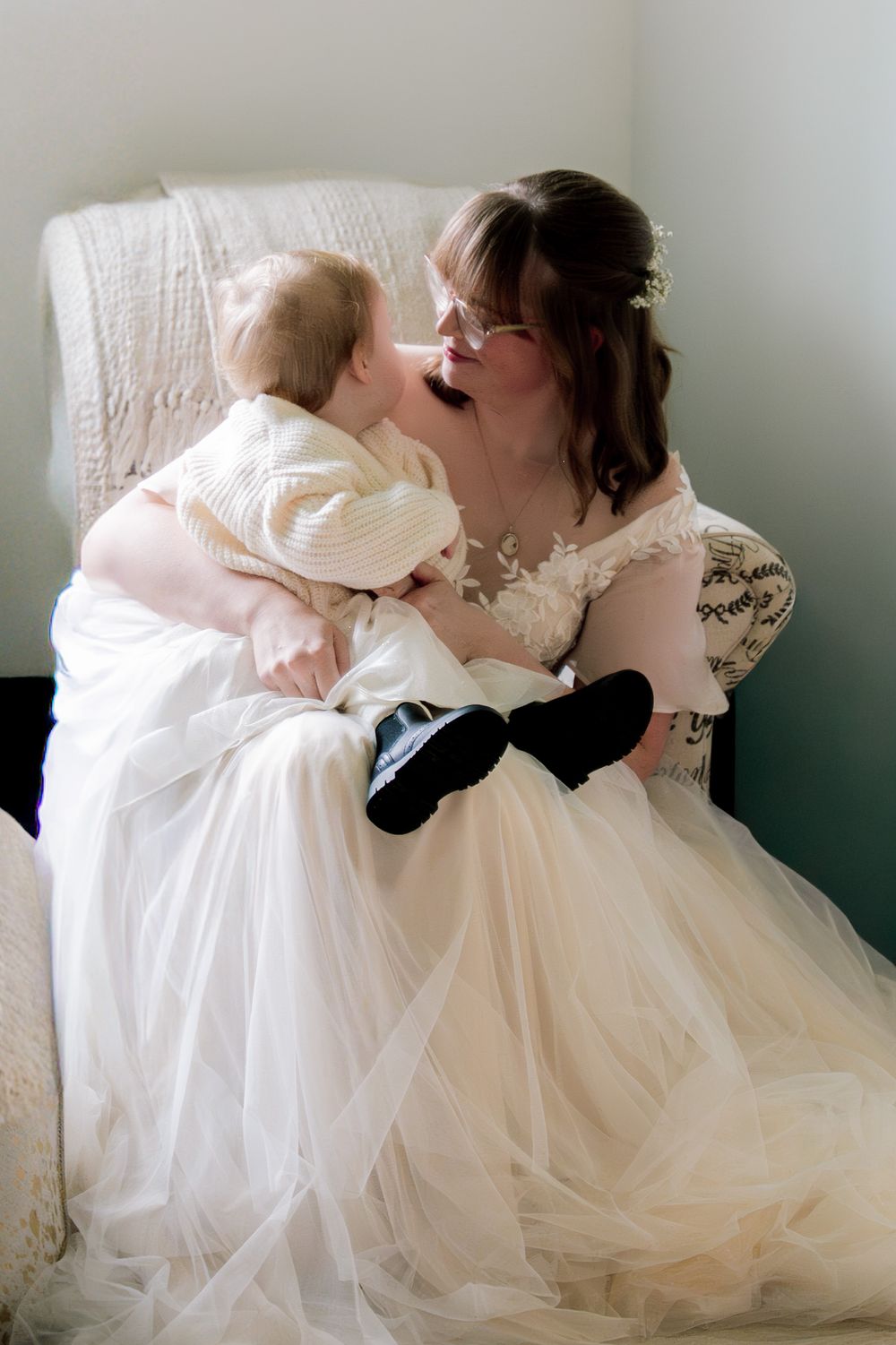 Bride in white lace gown sits in chair, lovingly holding and nuzzling a baby in cream outfit.