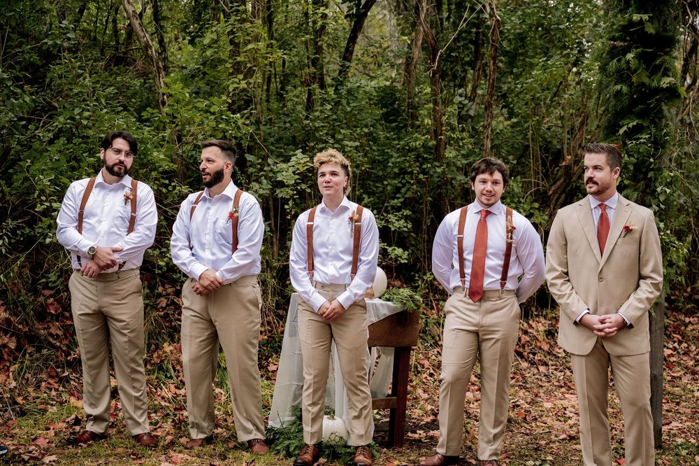 Five groomsmen in khaki pants, white shirts, and suspenders stand outdoors in a lush green forest setting.